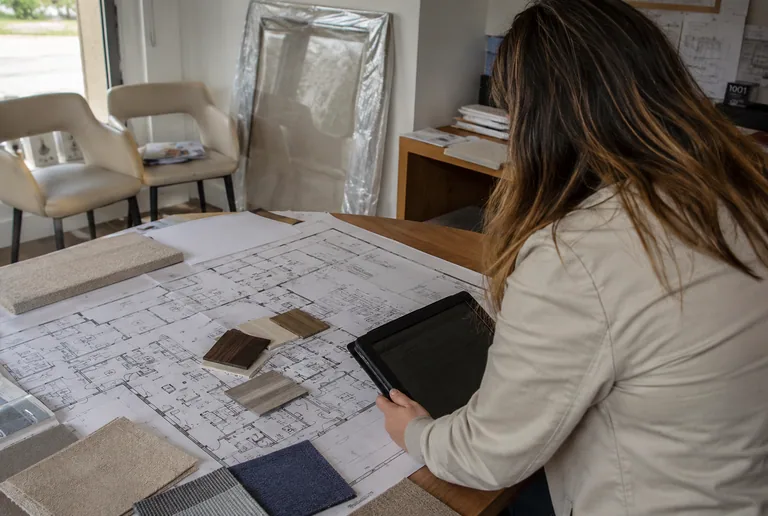 A designer leaning over printed floor plans covered in fabric and finish samples, holding a tablet, with sample chairs and a wrapped mirror behind.