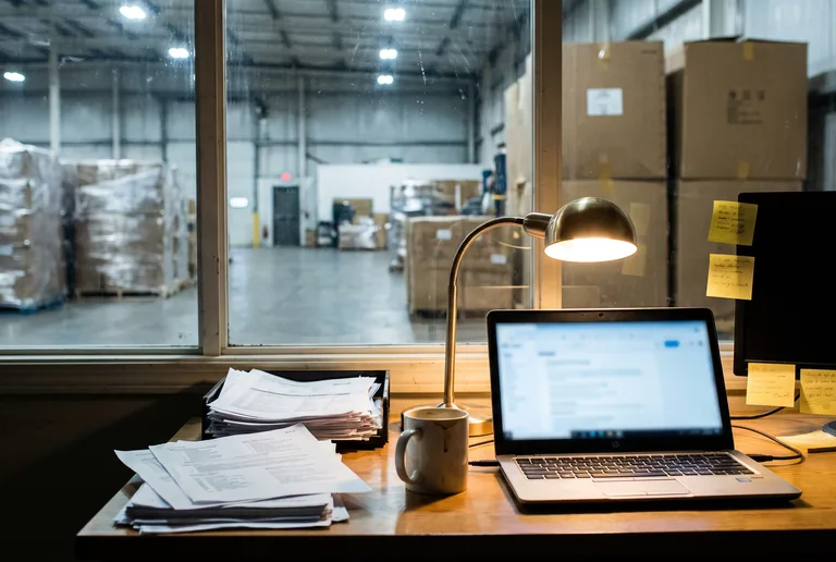 A warehouse office desk stacked with paperwork and an open laptop, overlooking a much larger furniture staging floor through an interior window.